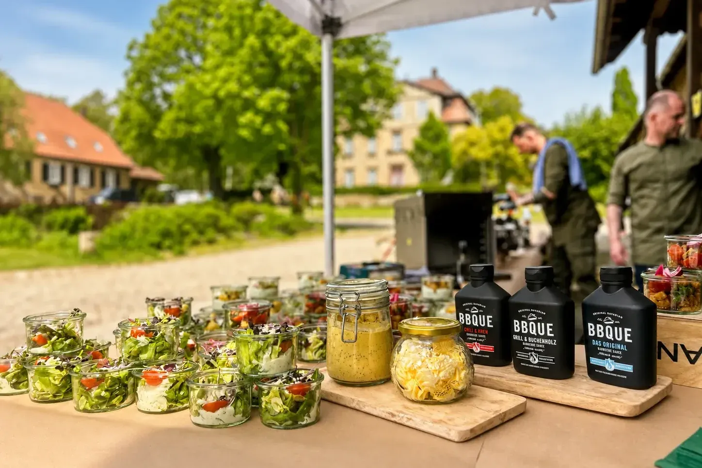 Catering-Tisch im Freien mit Salatgläsern und BBQ-Saucen, Grill im Hintergrund