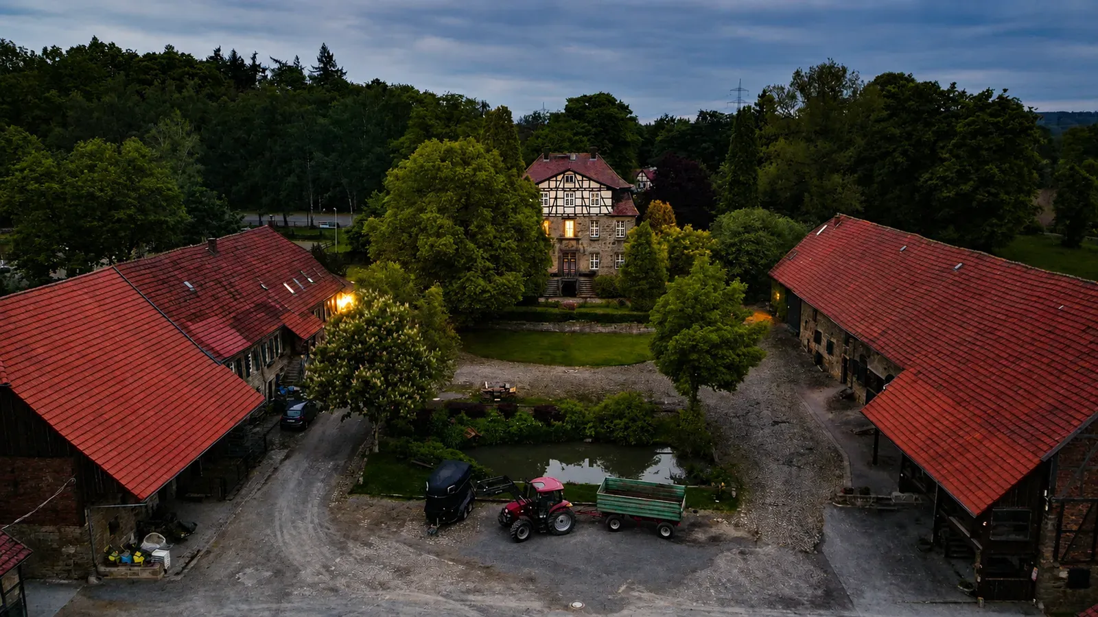 Gutshof in der Dämmerung mit Fachwerk-Gutshaus, zwei Scheunen und Traktor im Hof