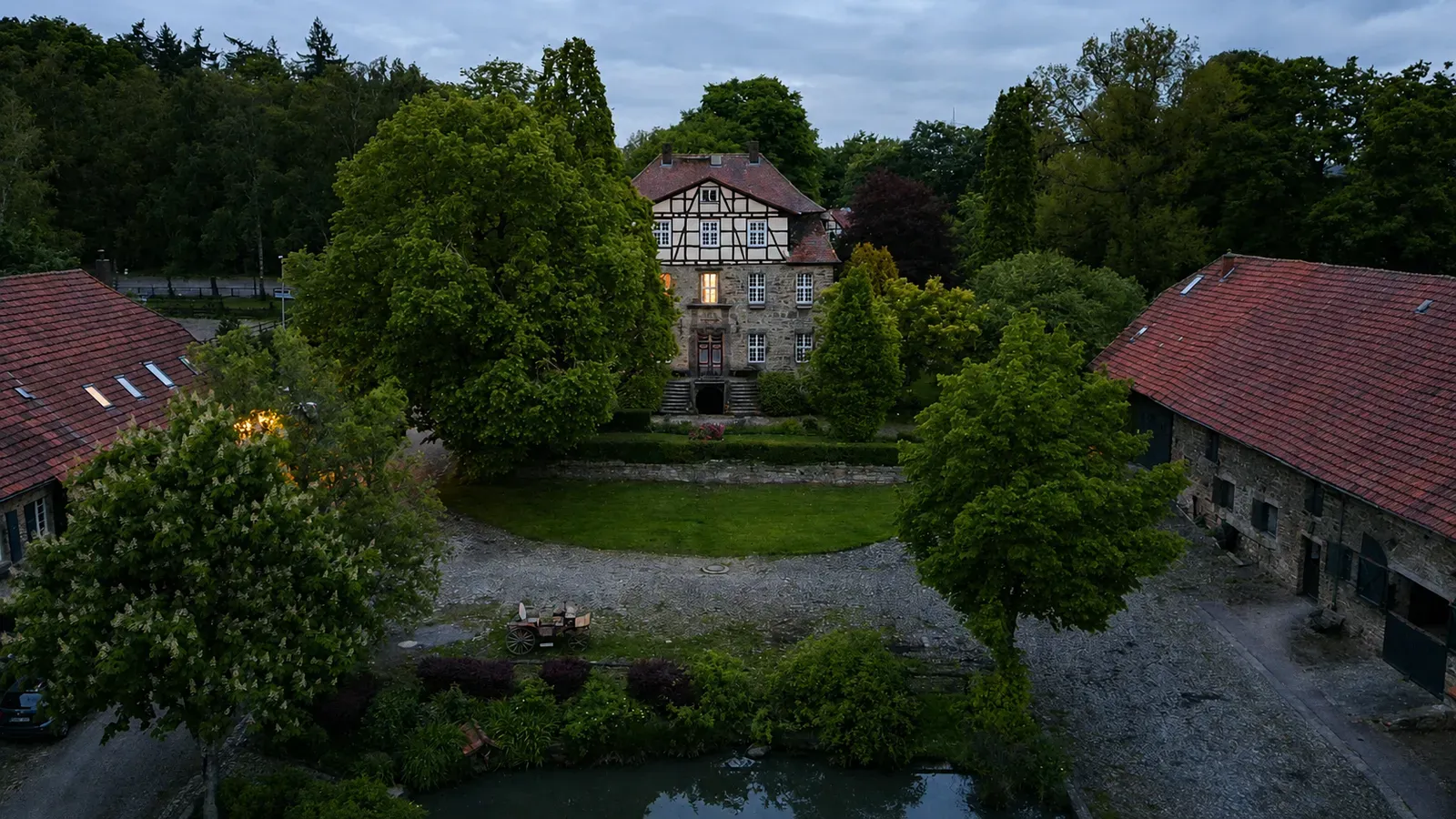 Fachwerk-Gutshaus hinter großen Bäumen mit Rasenhof, Teich und seitlichen Stallgebäuden